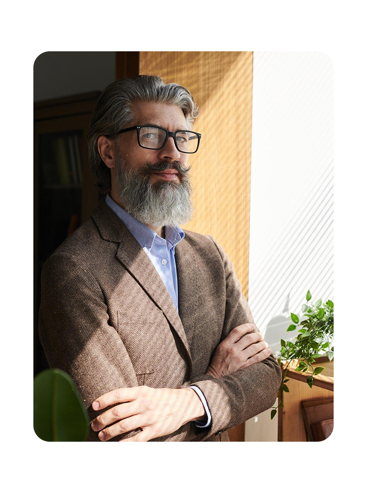 mature bearded man wearing stylish outfit and eyeglasses standing with arms crossed looking at camera